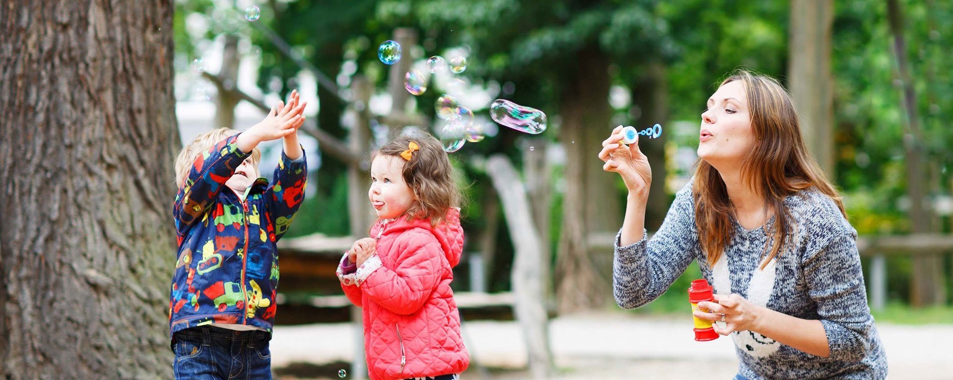 Zwei Kinder spielen mit ihrer Mama draußen und fangen Seifenblasen mit ihren Händen.